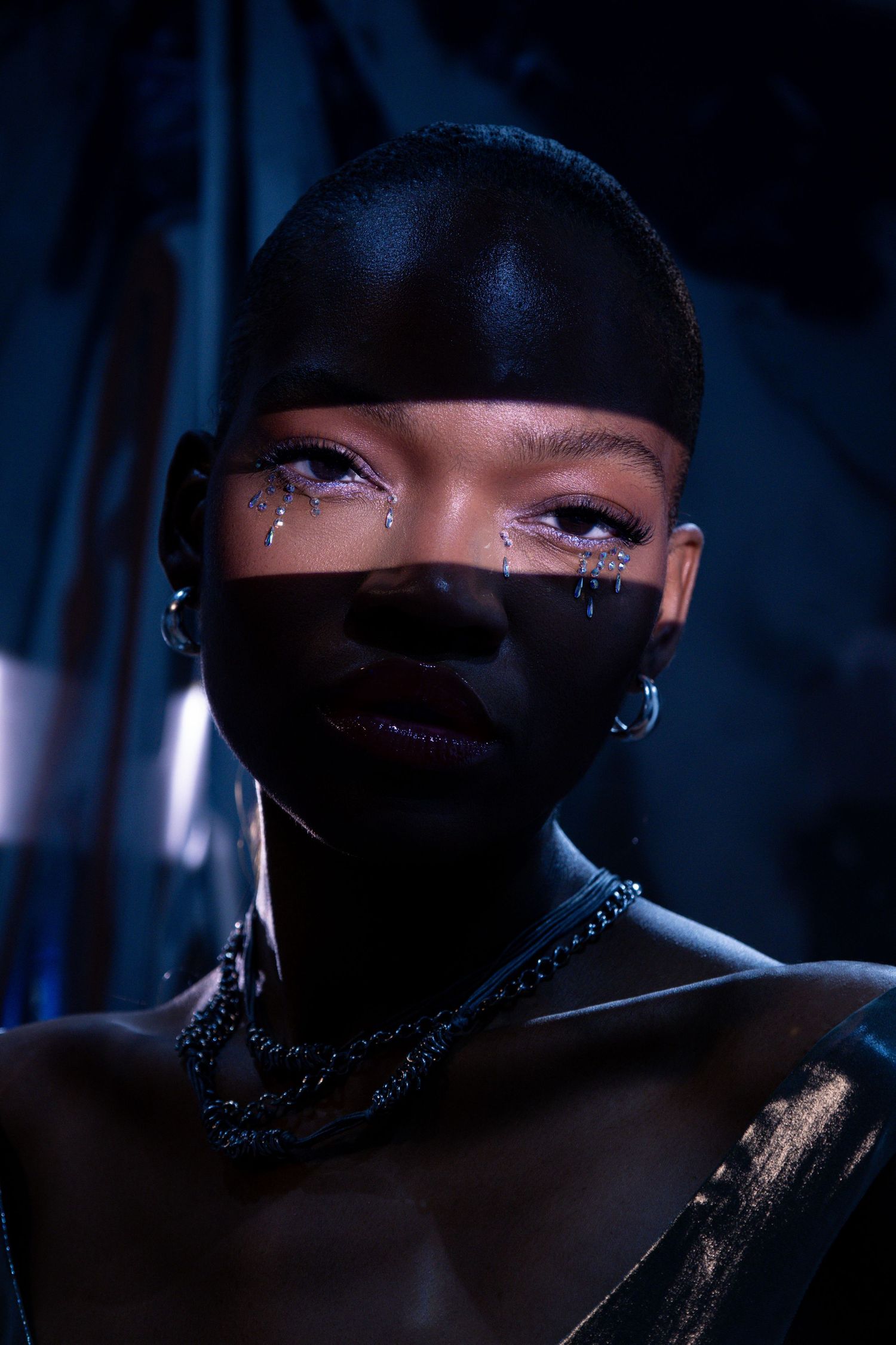 Black woman with artistic teardrop makeup, hoop earrings, and chain necklace poses dramatically in dark blue studio lighting.