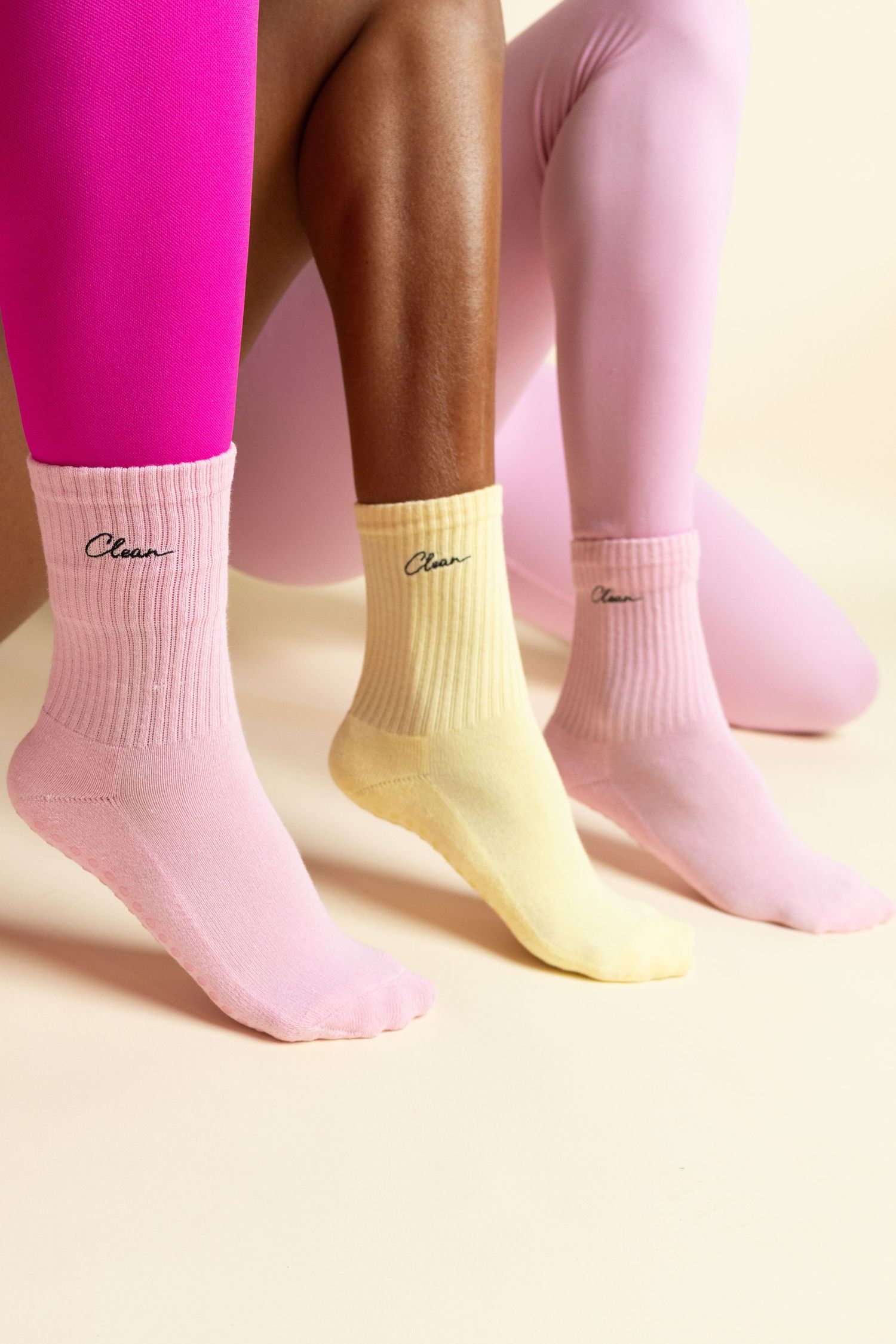 Three models wearing pink and yellow Clean-branded crew socks with grip soles, styled with colorful leggings.