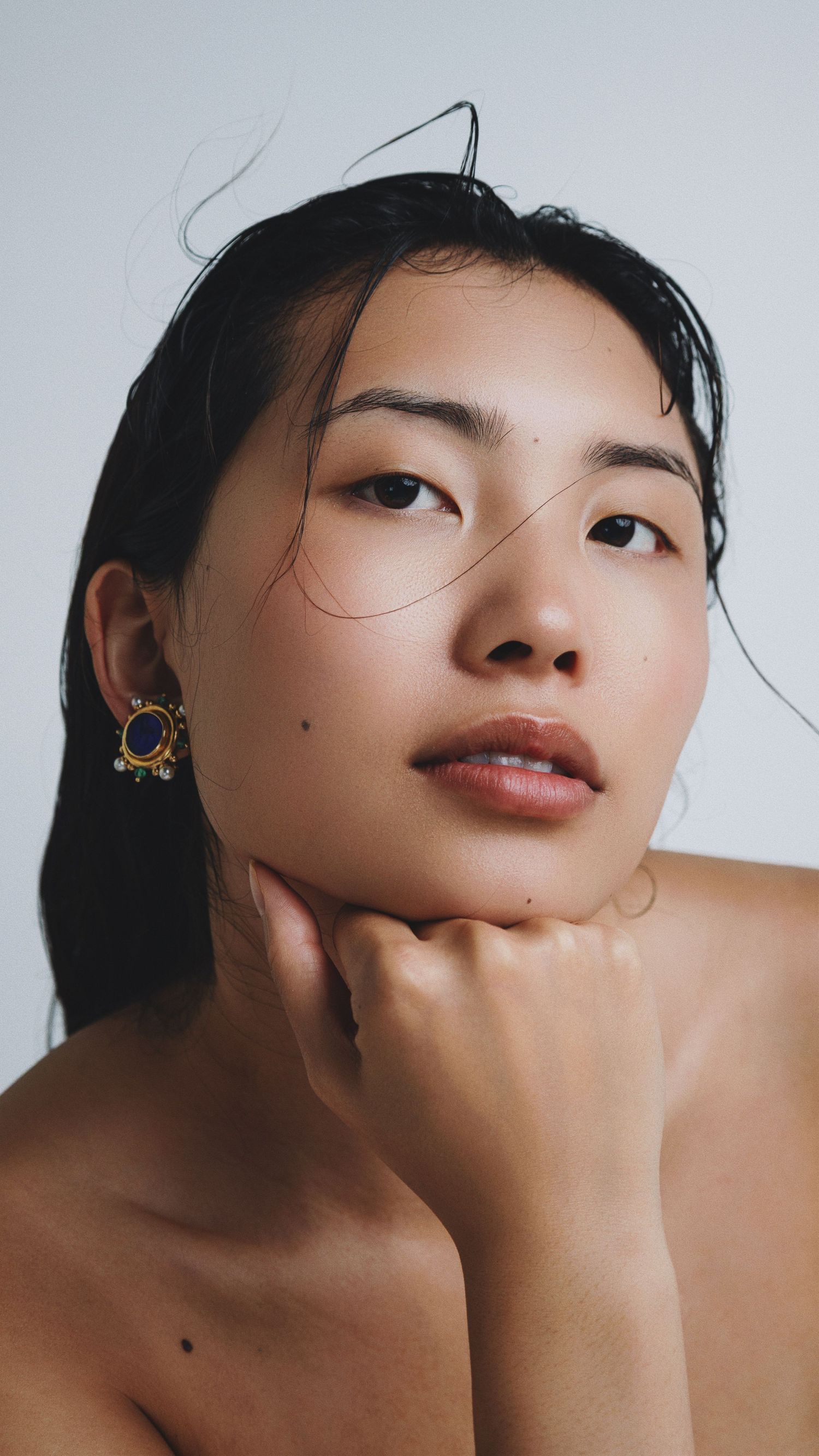 Young woman with dark hair rests chin on hand, wearing ornate gold and gemstone earrings against a light background.