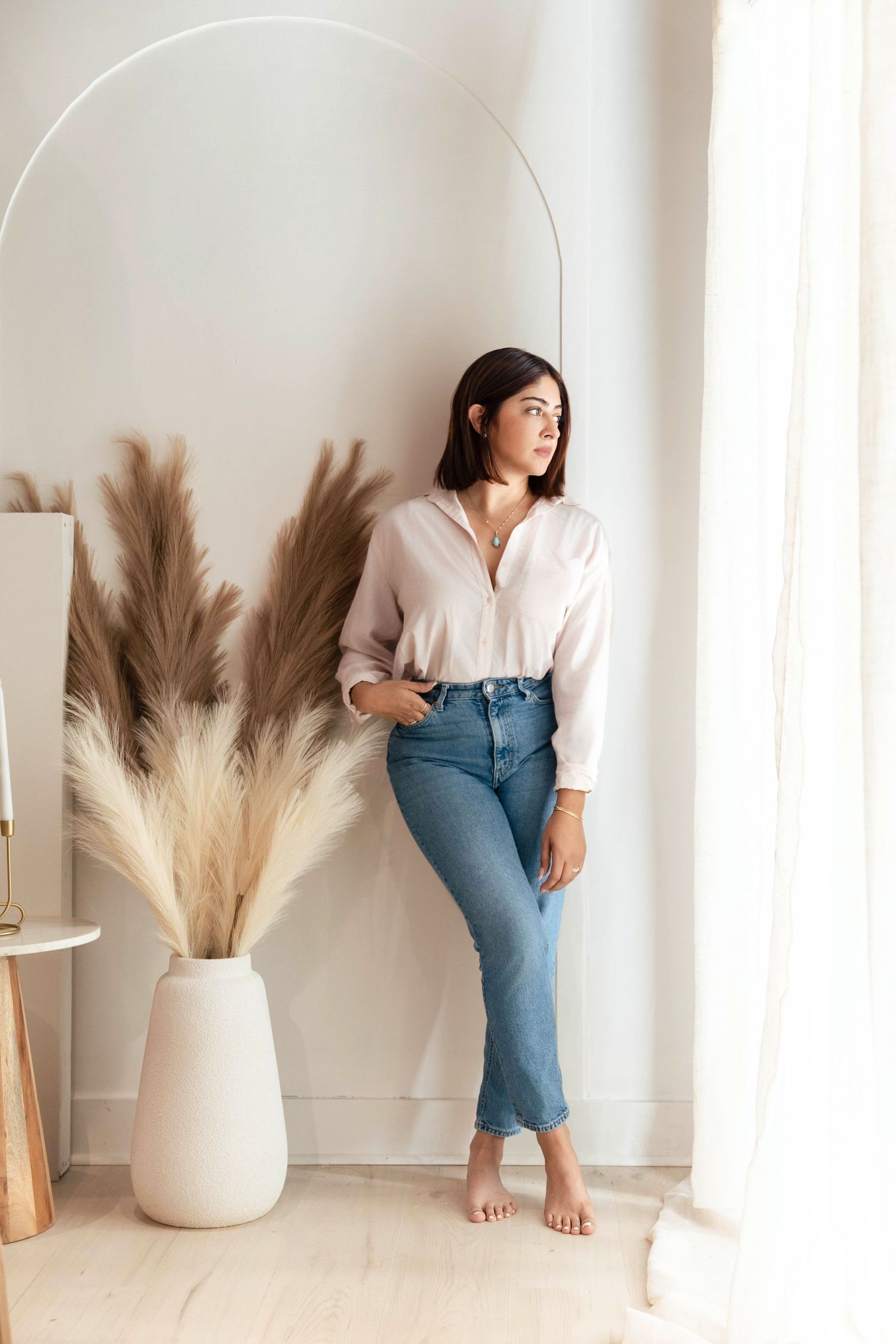 Woman in white blouse and blue jeans leaning against wall near pampas grass decor by sheer curtains.