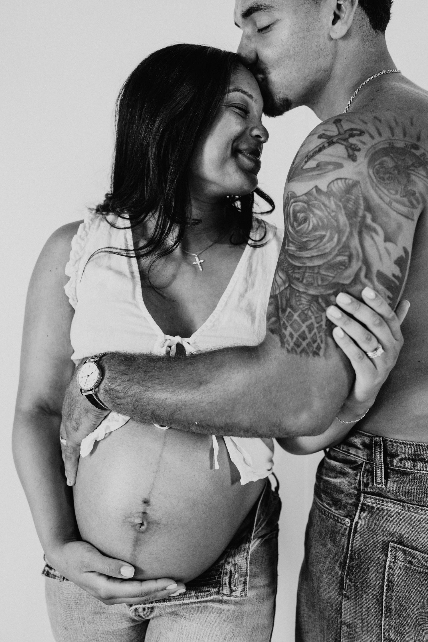 Black and white maternity photo of a pregnant woman being embraced and kissed by a tattooed man.