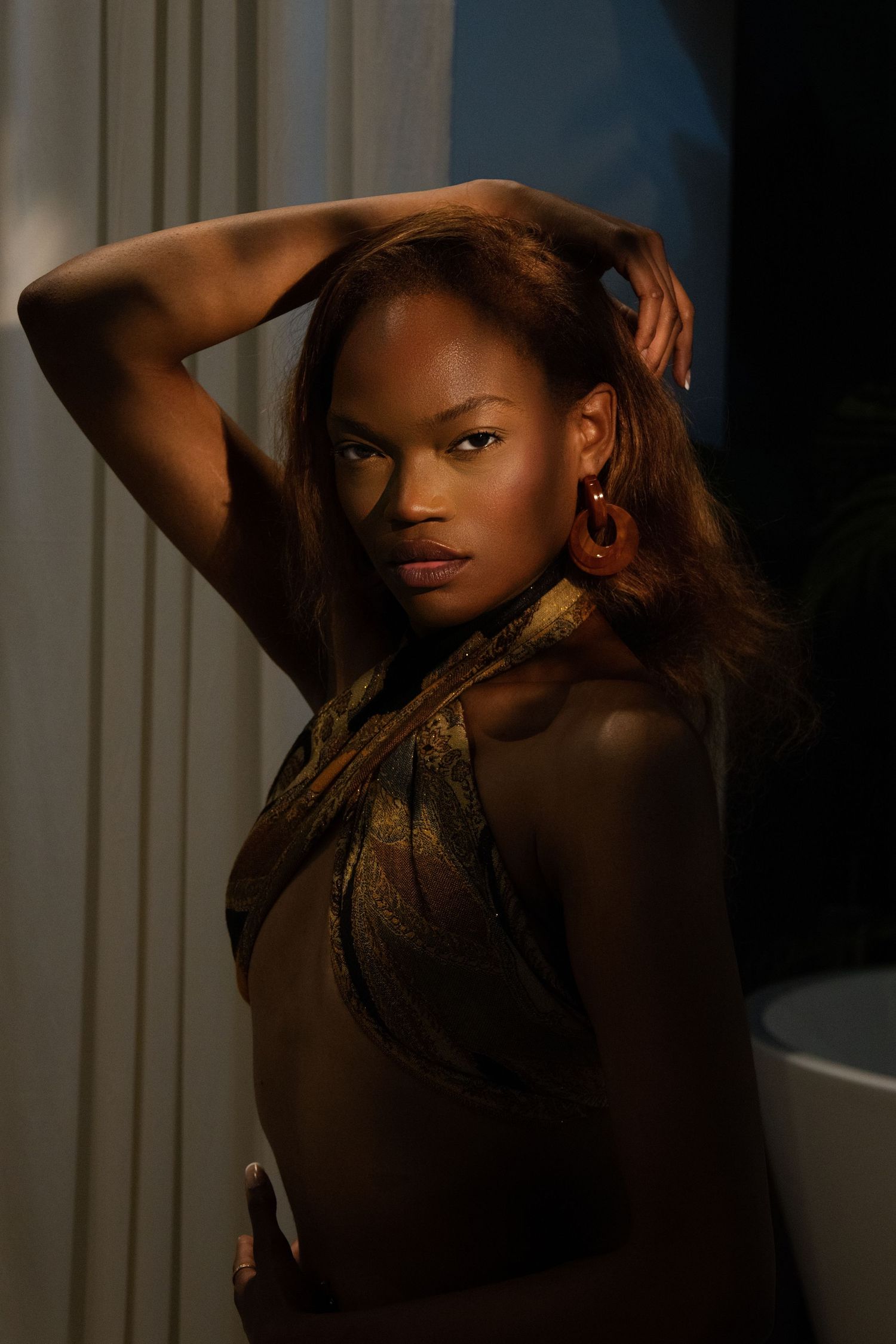 A Black woman poses confidently in dramatic lighting, wearing a gold halter top with hoop earrings, hands raised behind her head.