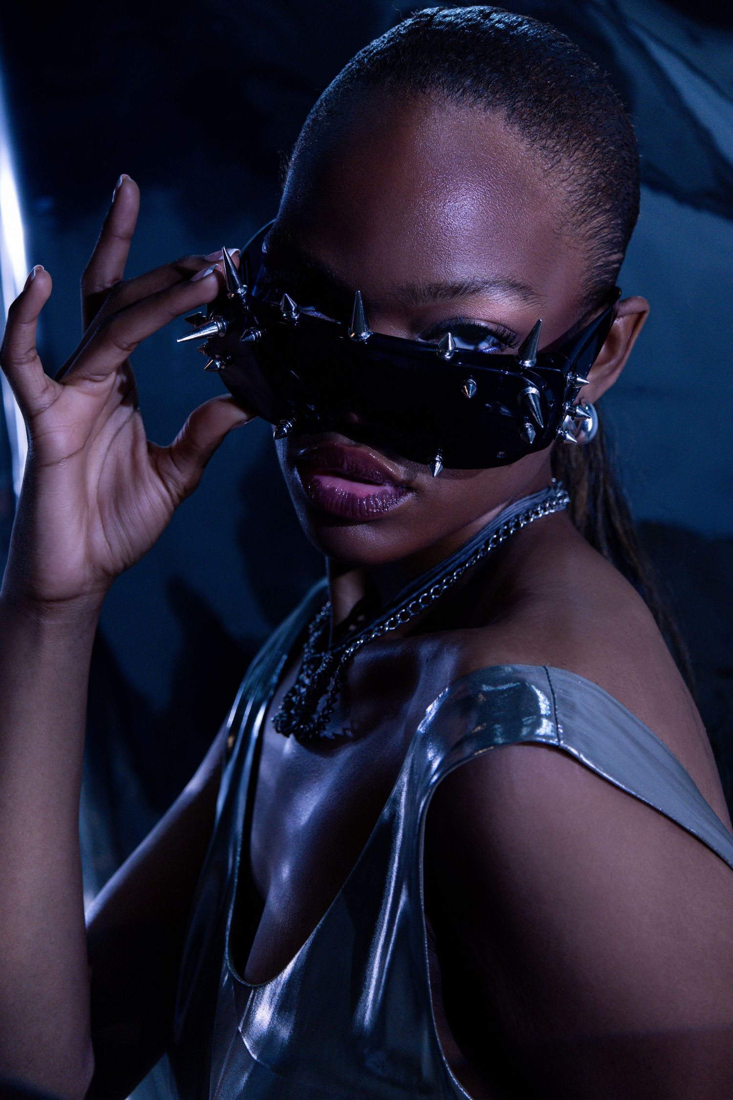 Woman wearing spiked black sunglasses and silver metallic top poses dramatically in dark studio setting.