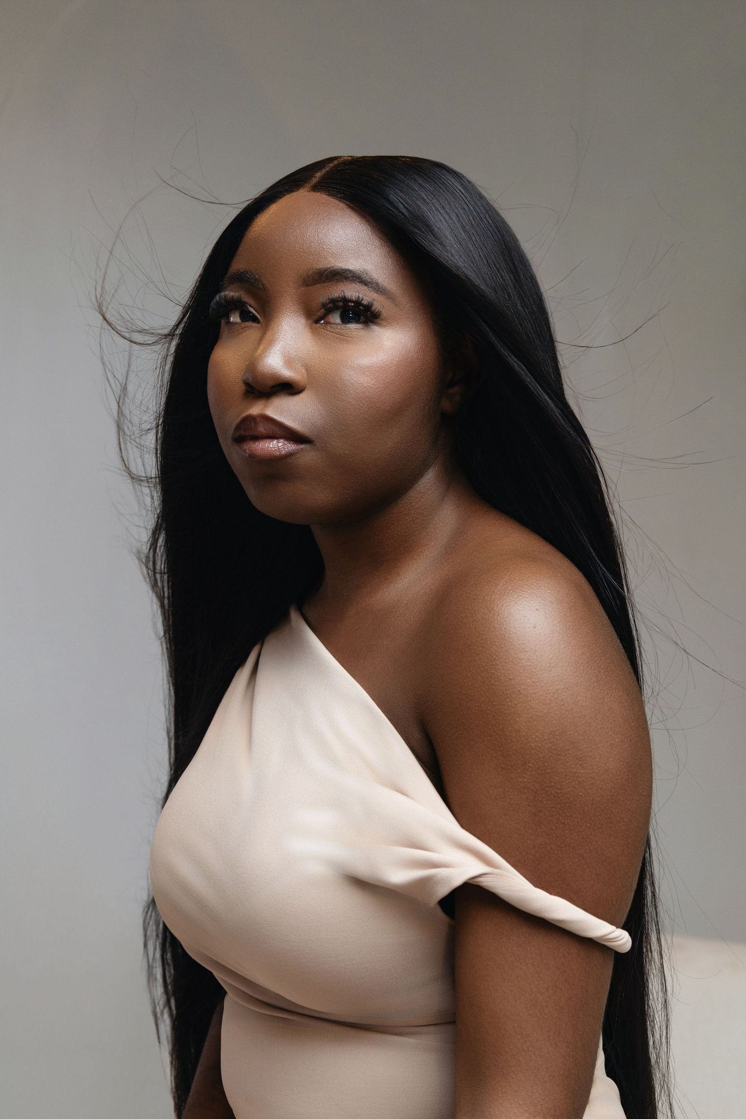 A Black woman with long straight hair wearing a beige off-shoulder top poses gracefully against a neutral background.