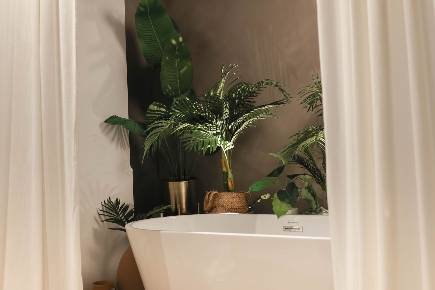 Freestanding white bathtub surrounded by lush tropical plants in a modern bathroom with sheer curtains.