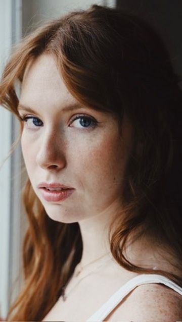 Young woman with red hair, blue eyes, and freckles gazing thoughtfully near a window in natural light.