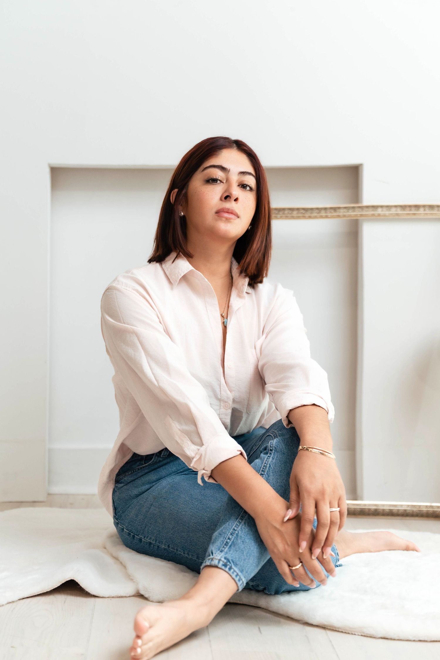 A young woman in a white shirt and jeans sits cross-legged on a white rug against a light wall.
