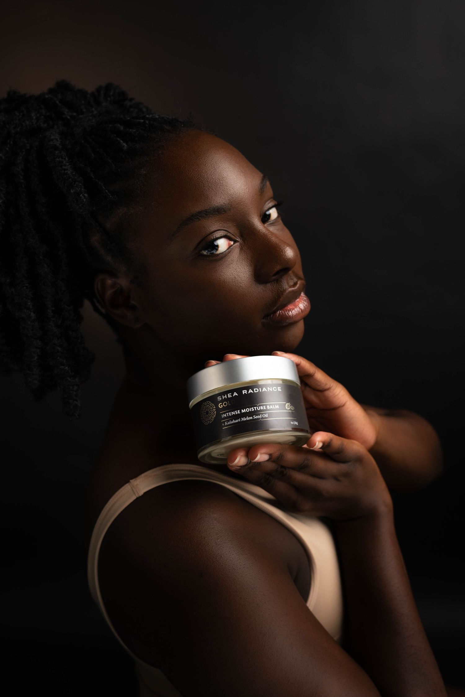 Young woman with braided hair holds a Shea Radiance skincare jar against dark background, showcasing glowing skin.