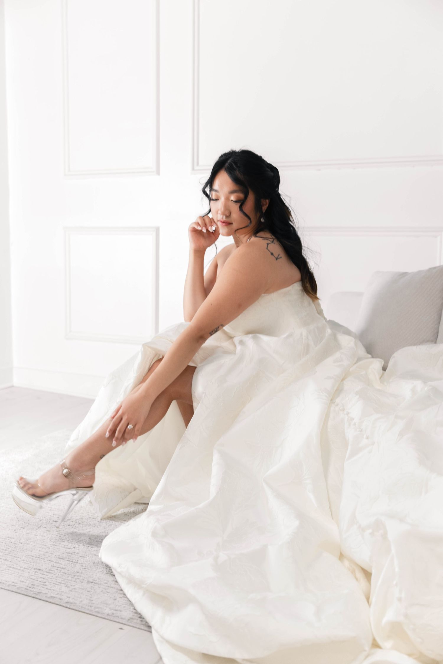 Elegant bride in a white wedding gown sitting on the floor, wearing heels, with dark hair and a shoulder tattoo.