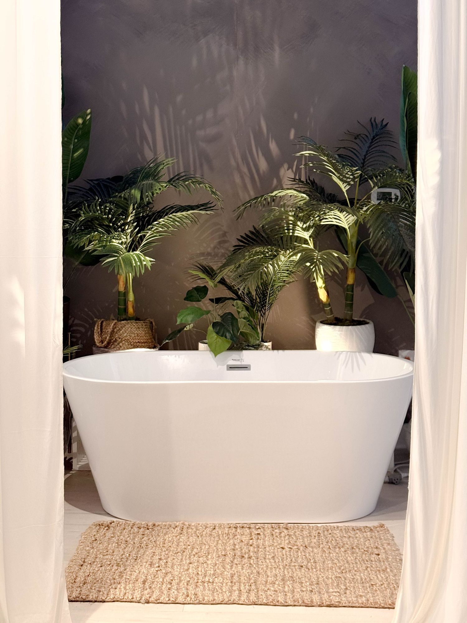 White freestanding bathtub surrounded by lush tropical plants on a jute rug in a serene, minimalist bathroom.