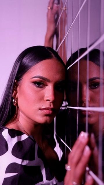 Woman with sleek black hair and gold earrings leans against mirrored blinds in purple-lit artistic portrait.