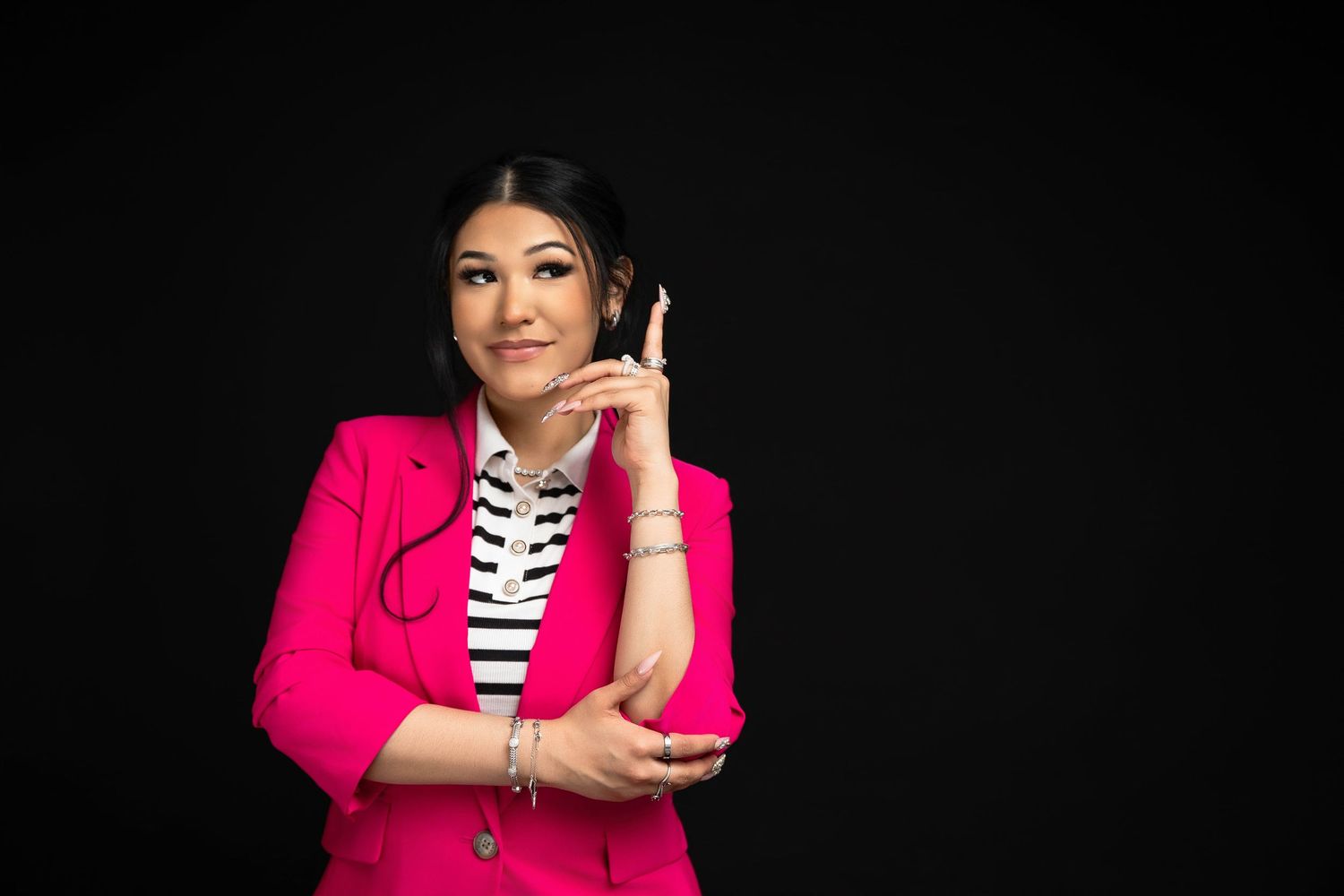 Young Asian woman in pink blazer talking on phone, smiling thoughtfully against black background.