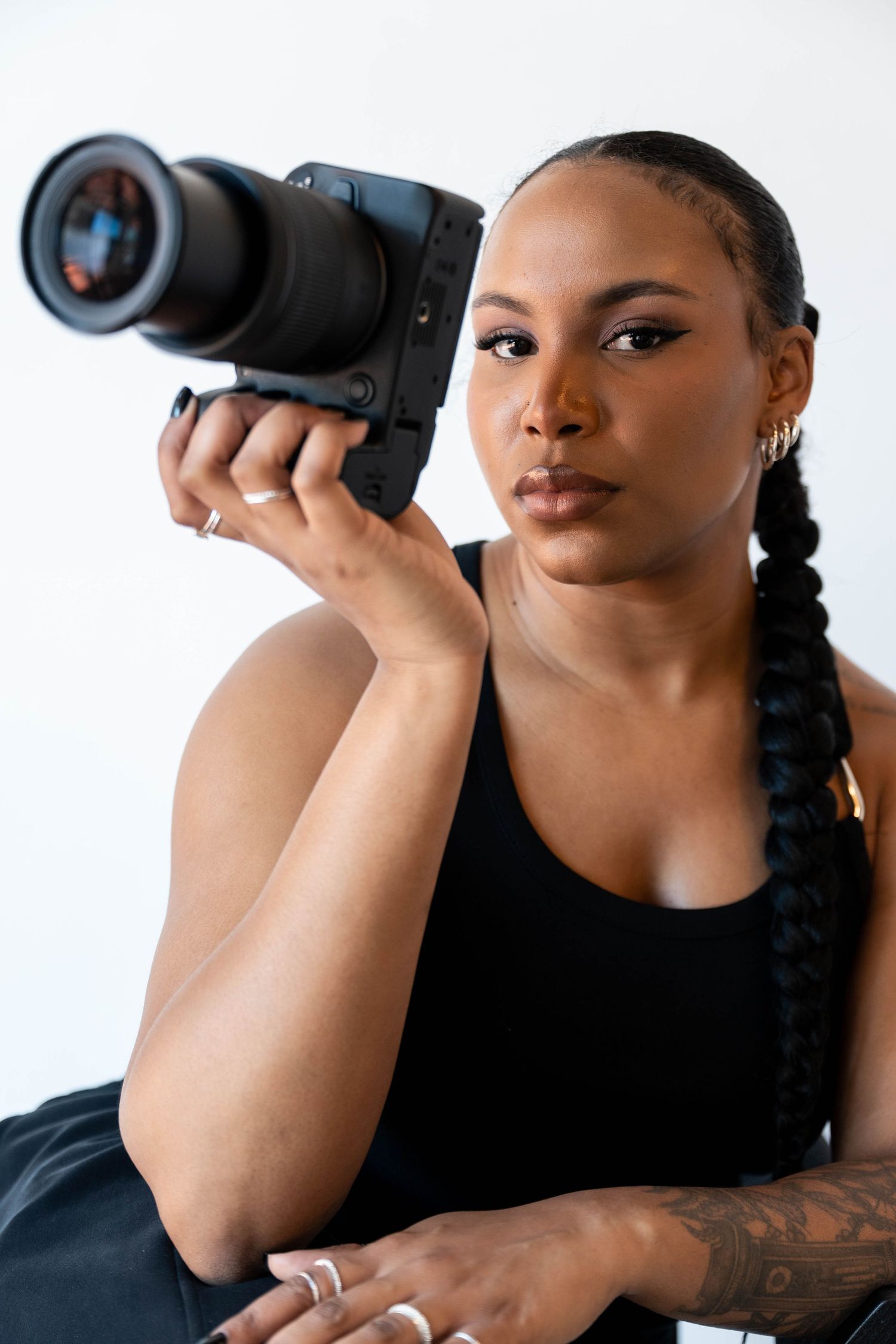Woman with braids holds professional camera with telephoto lens, wearing black tank top with tattoo visible on arm.