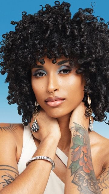 Woman with voluminous black curly hair, tattoos, and statement earrings posing against a blue background.