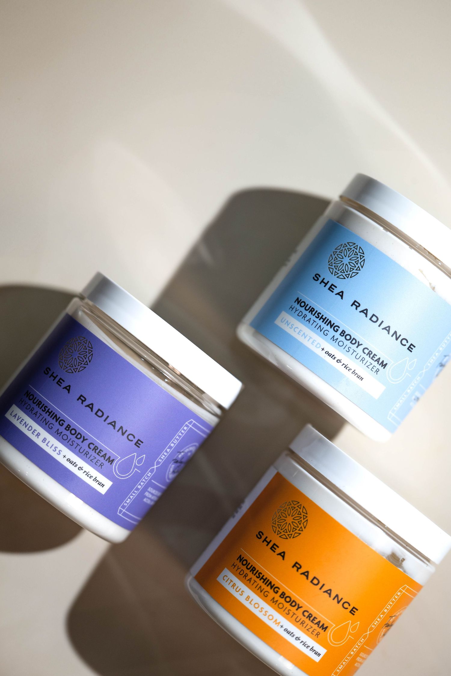 Three Shea Radiance nourishing body cream jars in purple, blue, and orange on a beige surface.