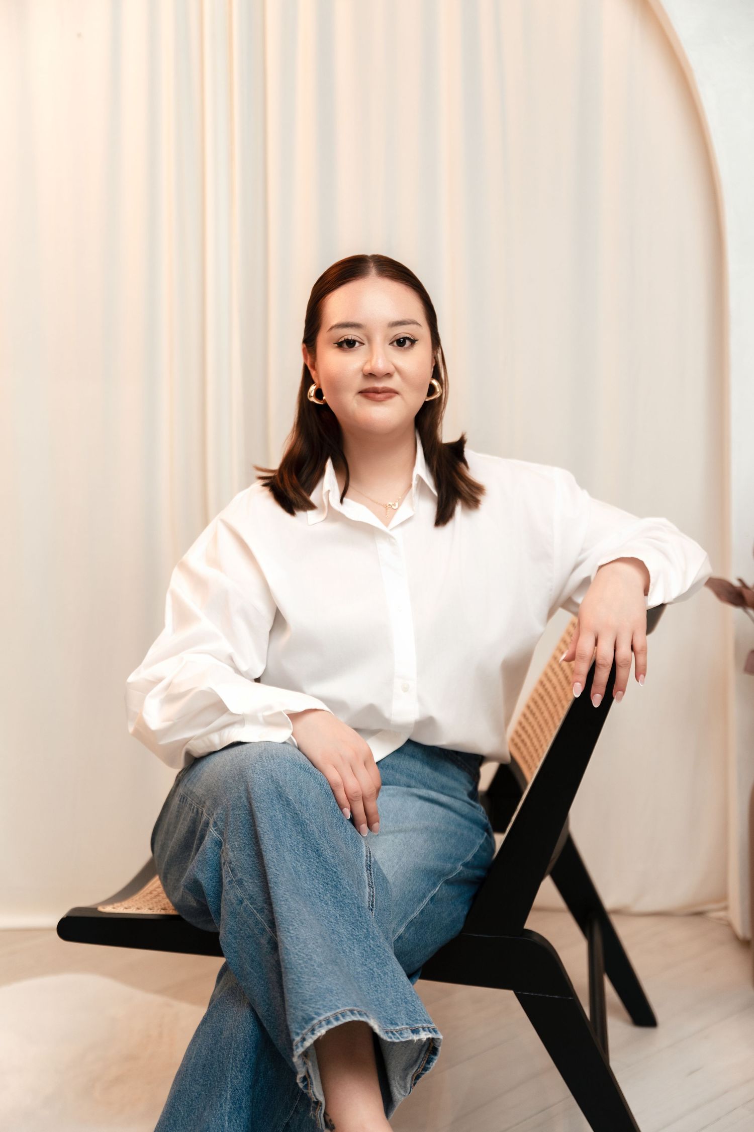 Young Asian woman in white shirt and jeans sitting relaxed on a modern chair against neutral curtain backdrop.