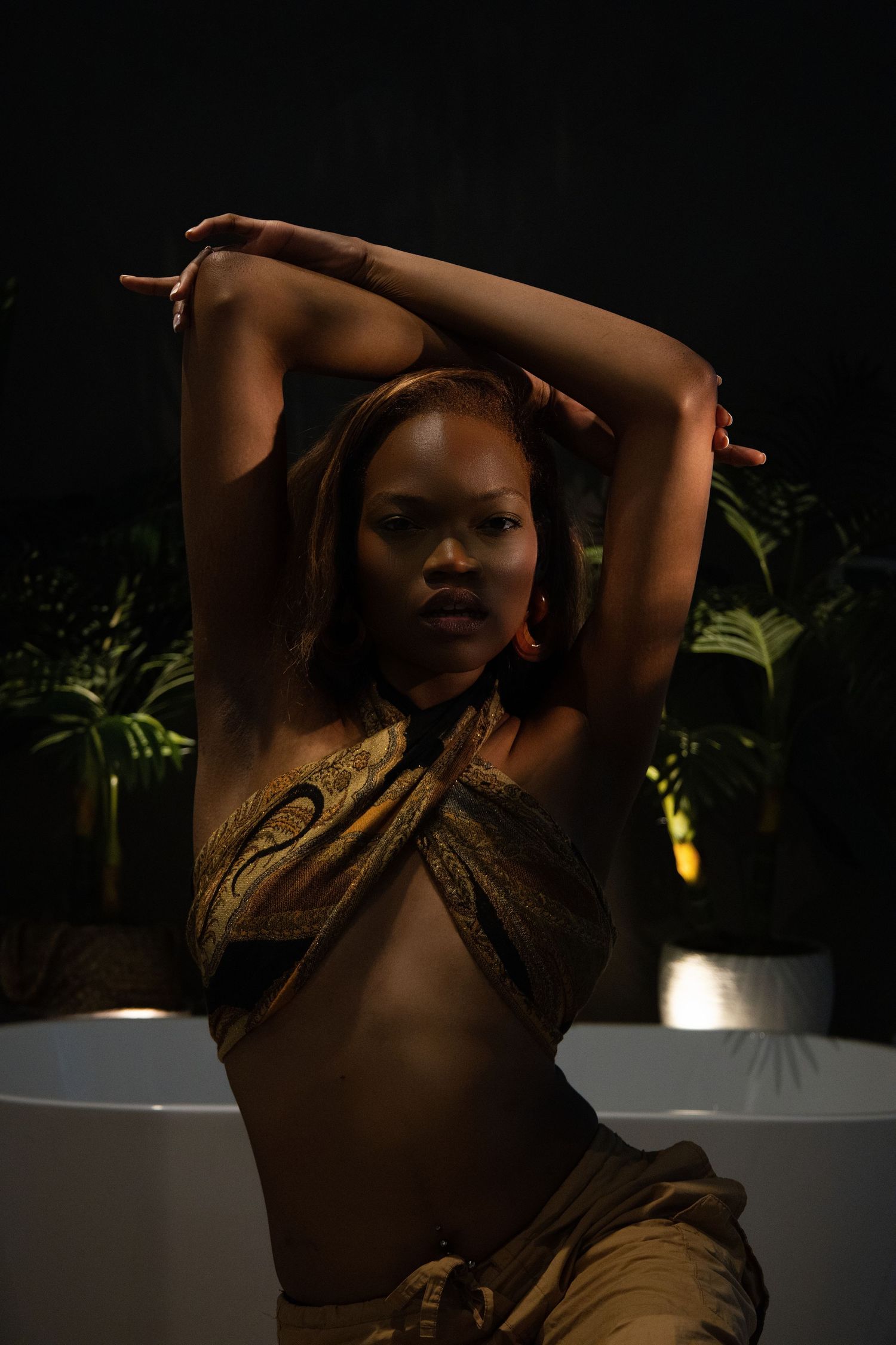 A woman in a golden wrap poses gracefully with arms raised against a dark background with tropical plants.