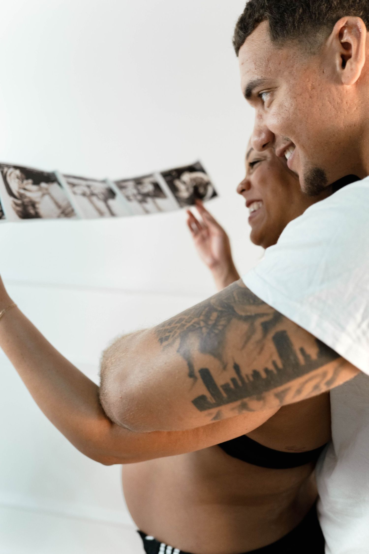 Pregnant couple smiling while reviewing ultrasound photos, man with city skyline tattoo embracing woman.