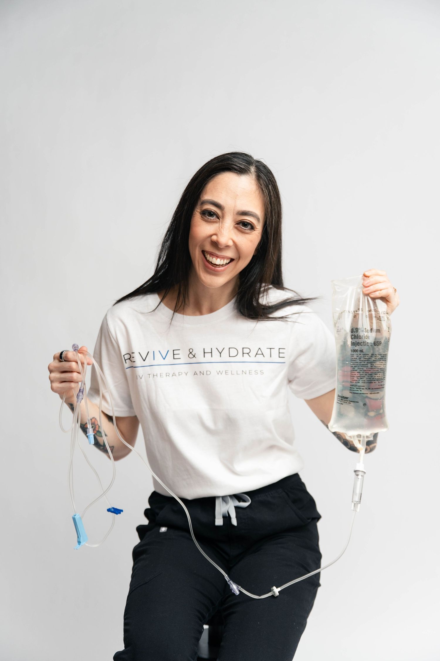 Smiling woman in Revive & Hydrate t-shirt holding IV therapy bags against white background.