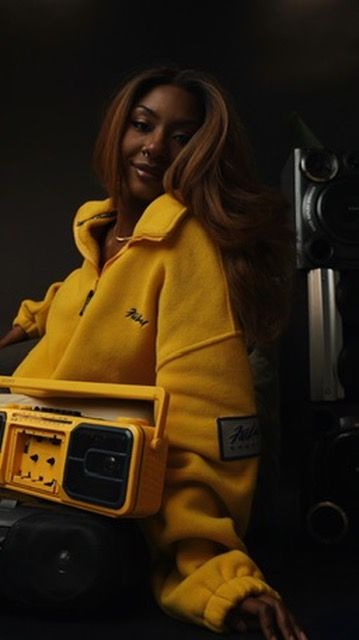Woman in yellow fleece jacket holding a matching yellow boombox in a dark studio setting.