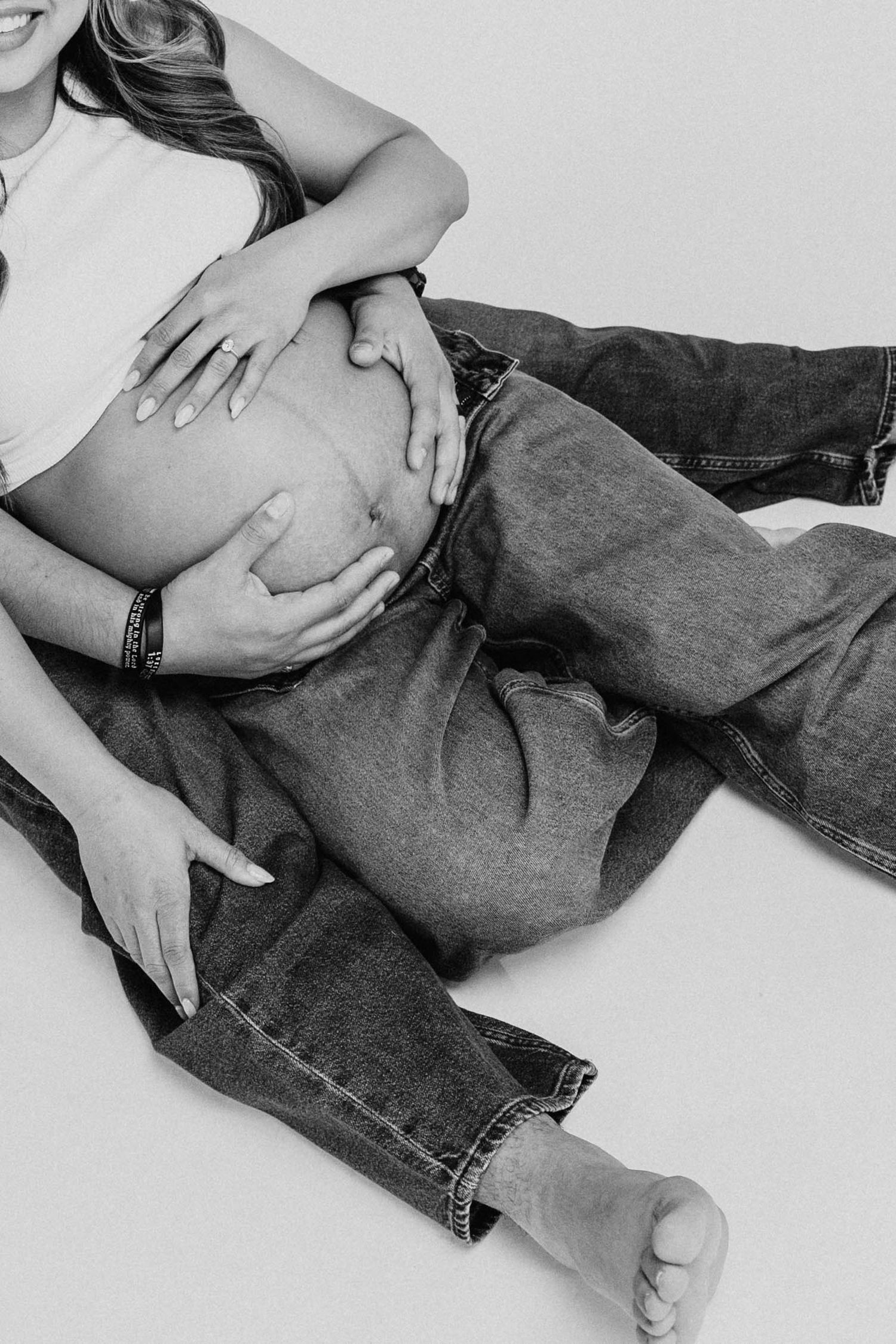 Black and white photo of a pregnant woman sitting on floor, cradling baby bump, wearing jeans and white crop top.