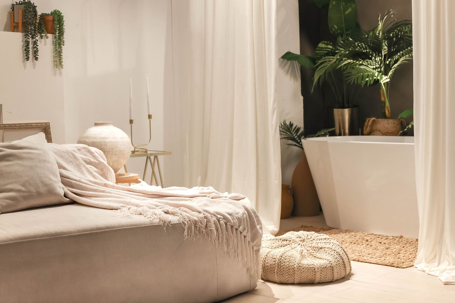Bright boho bedroom with neutral linen bedding, sheer curtains, woven pouf, and lush green plants near a freestanding bathtub.
