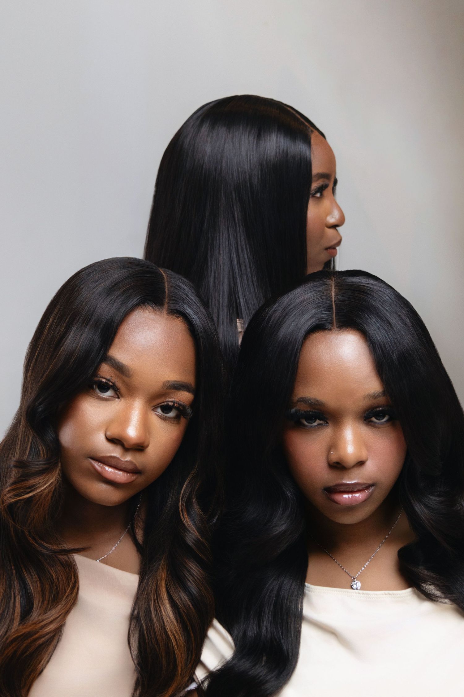 Three young Black women with long straight and wavy black hair pose together against a light gray background.