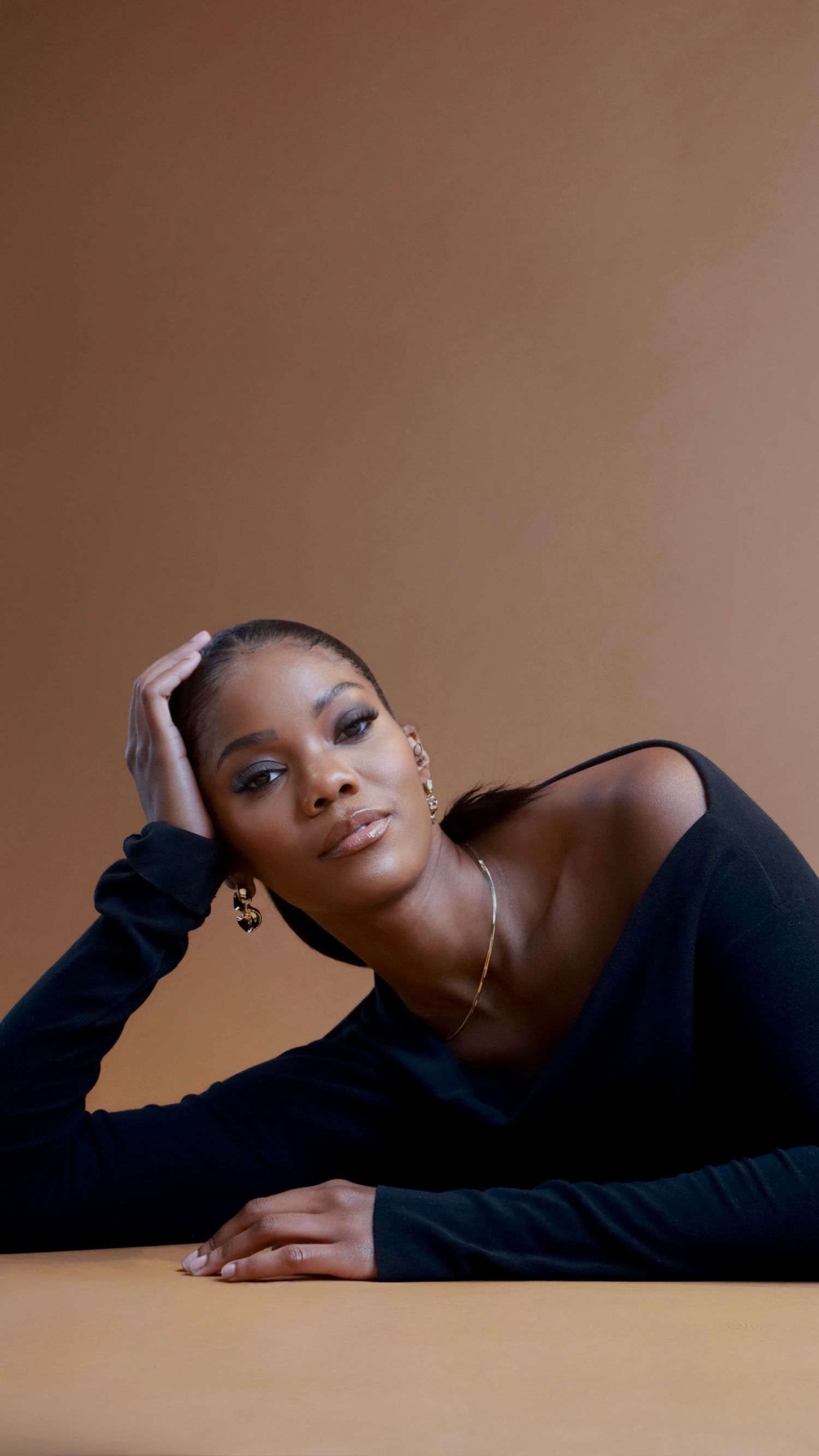 A woman in a black off-shoulder top poses elegantly at a table, wearing gold jewelry against a warm beige background.