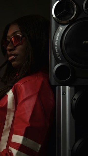 A woman in a red jacket and sunglasses poses next to a large bass reflex speaker in a dark setting.