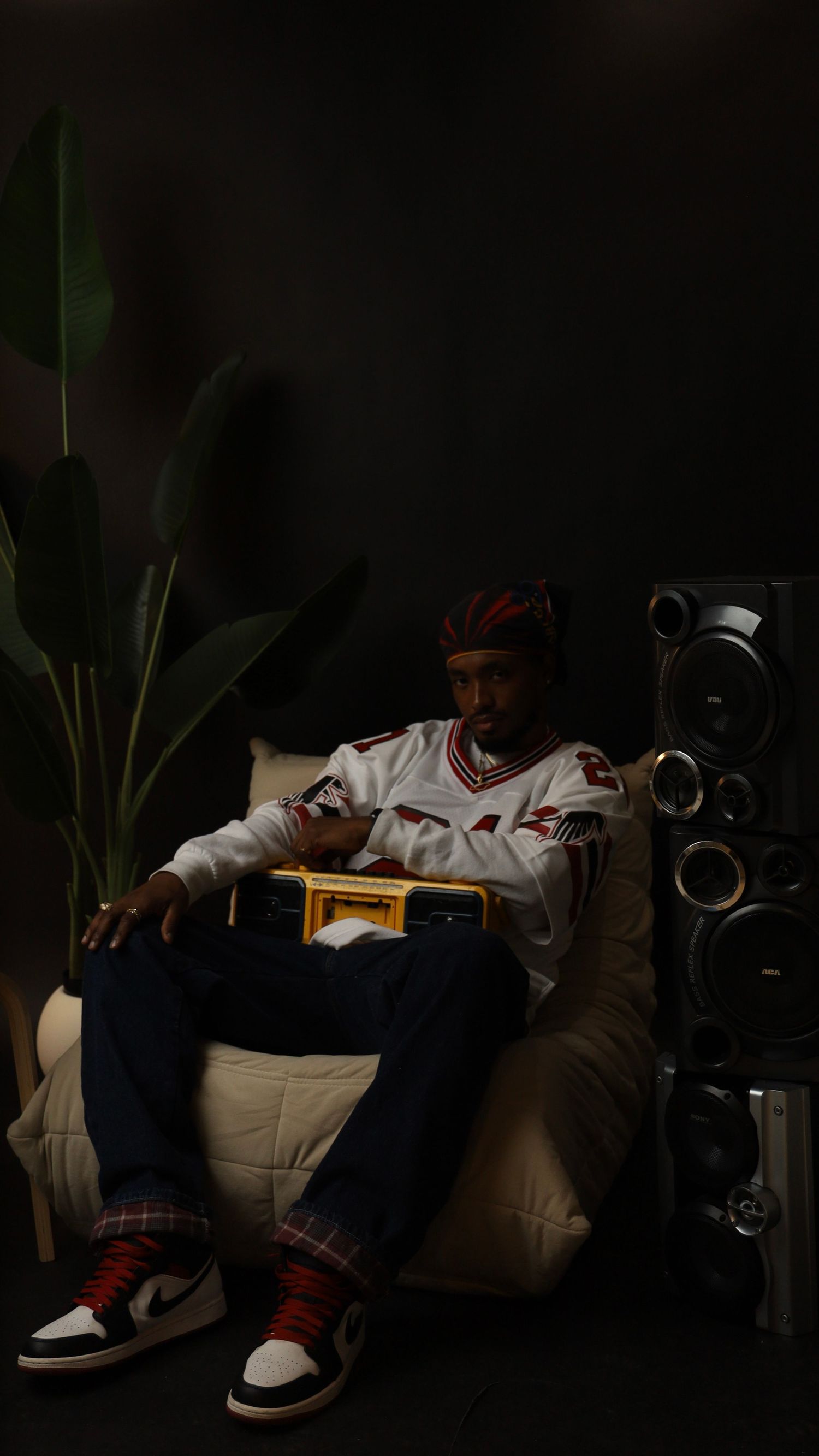Man in sports jersey and Air Jordan sneakers sits in bean bag chair holding yellow boombox beside speaker tower and plant.