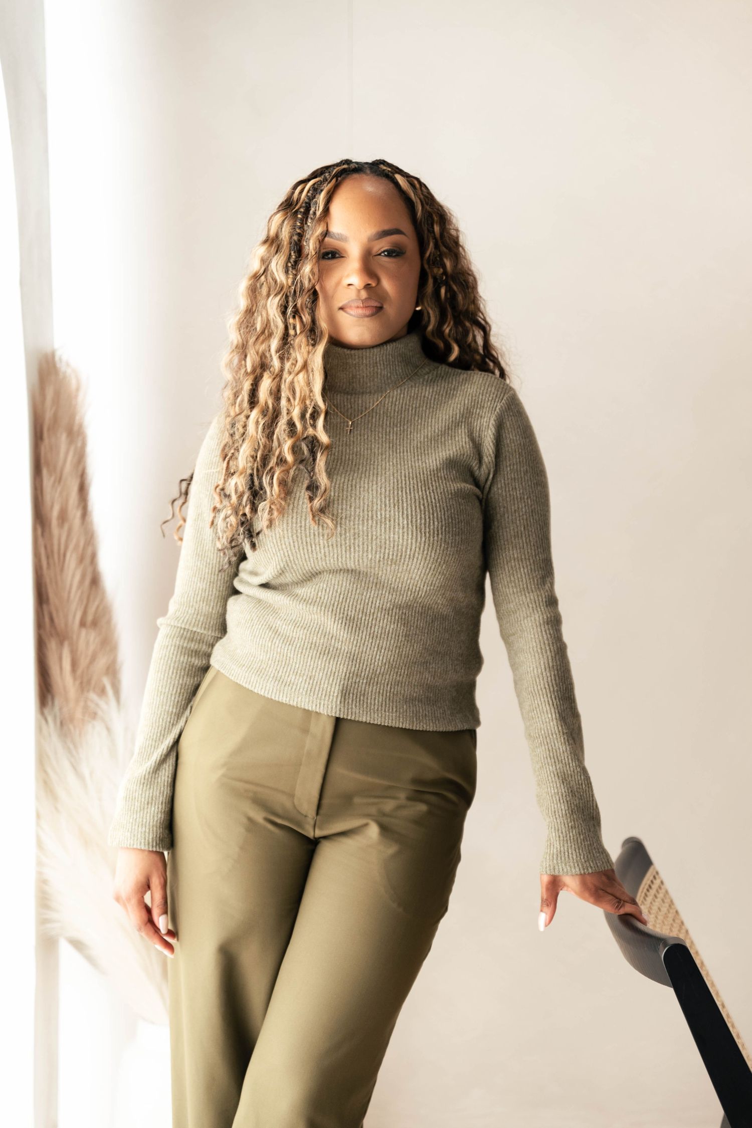 Woman in olive turtleneck sweater and matching trousers posing confidently against a white background.