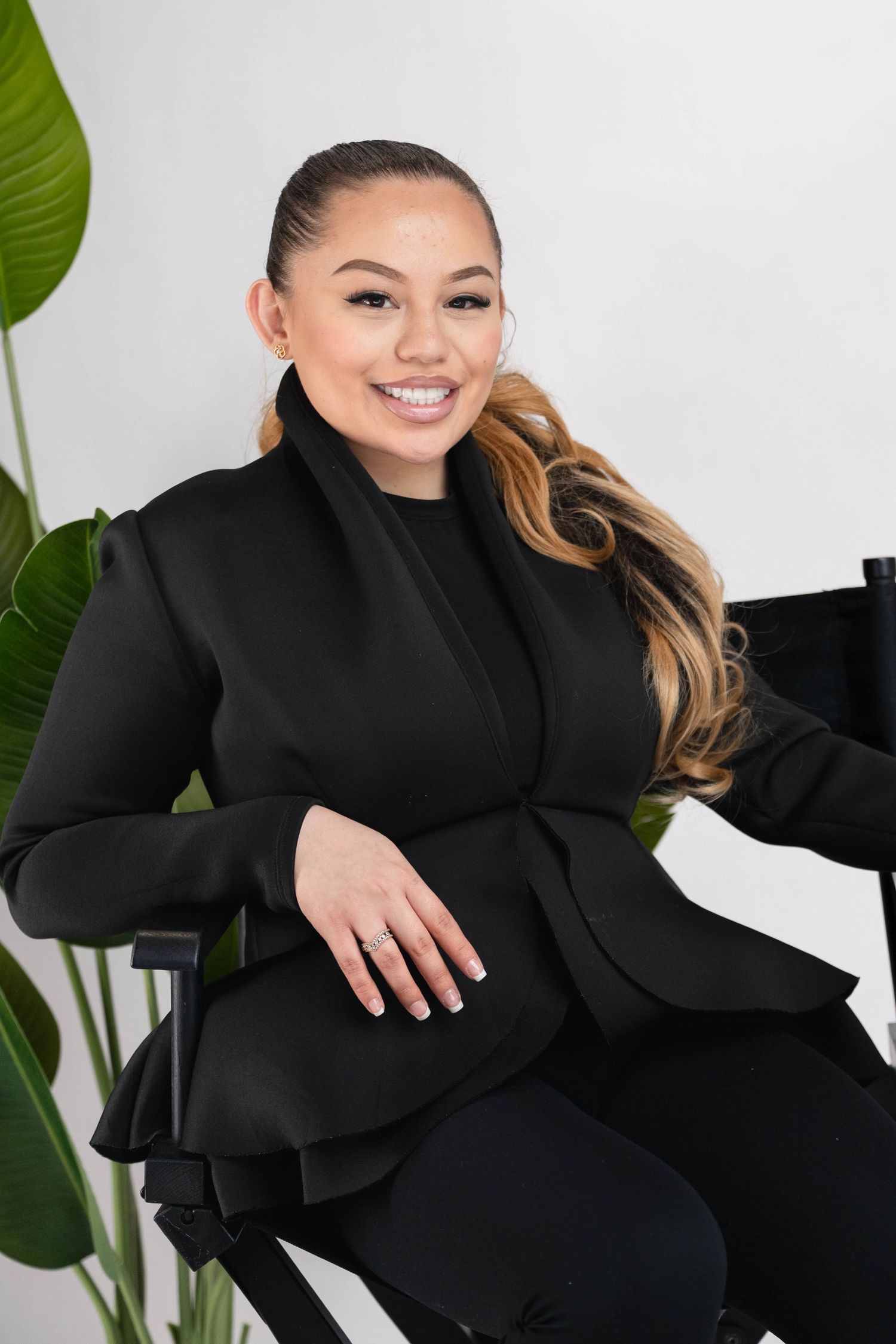 Smiling woman in black blazer seated in director chair with green tropical plant background.