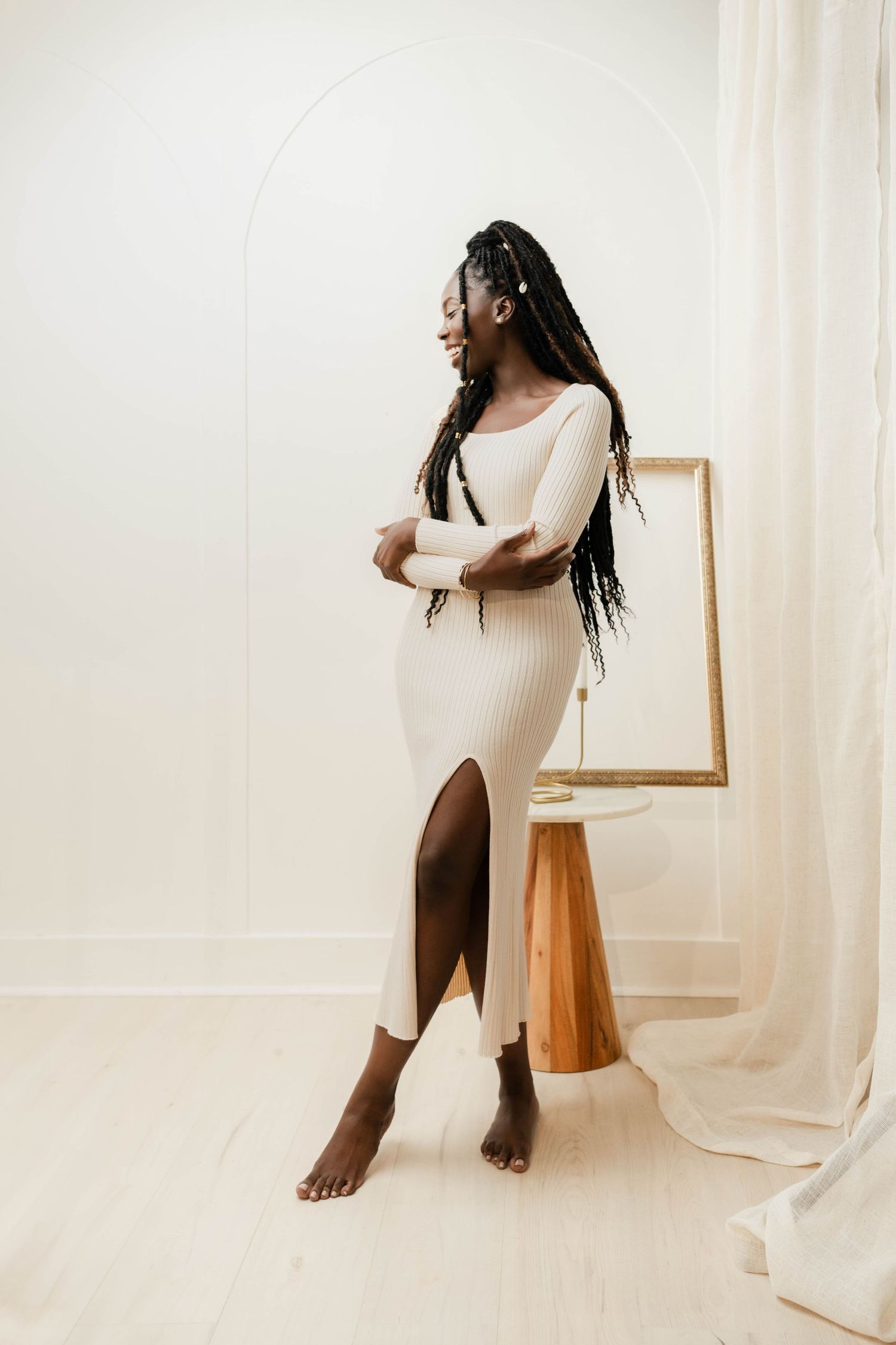 Black woman in cream ribbed midi dress with slit, long braids, posing barefoot in minimalist white studio.