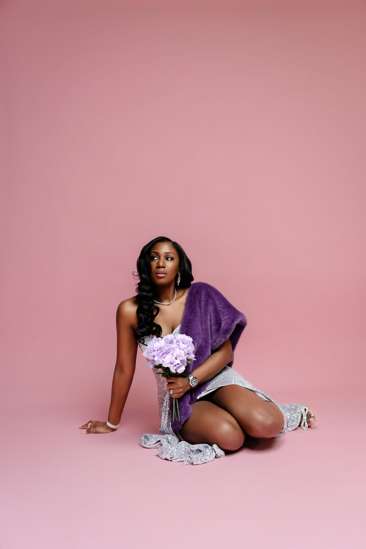 A woman in purple wrap and denim shorts sits on pink background holding lavender flowers.