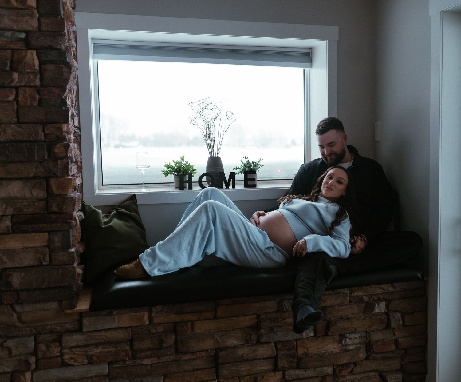 Expecting couple relaxes on stone window seat with HOME sign decor.
