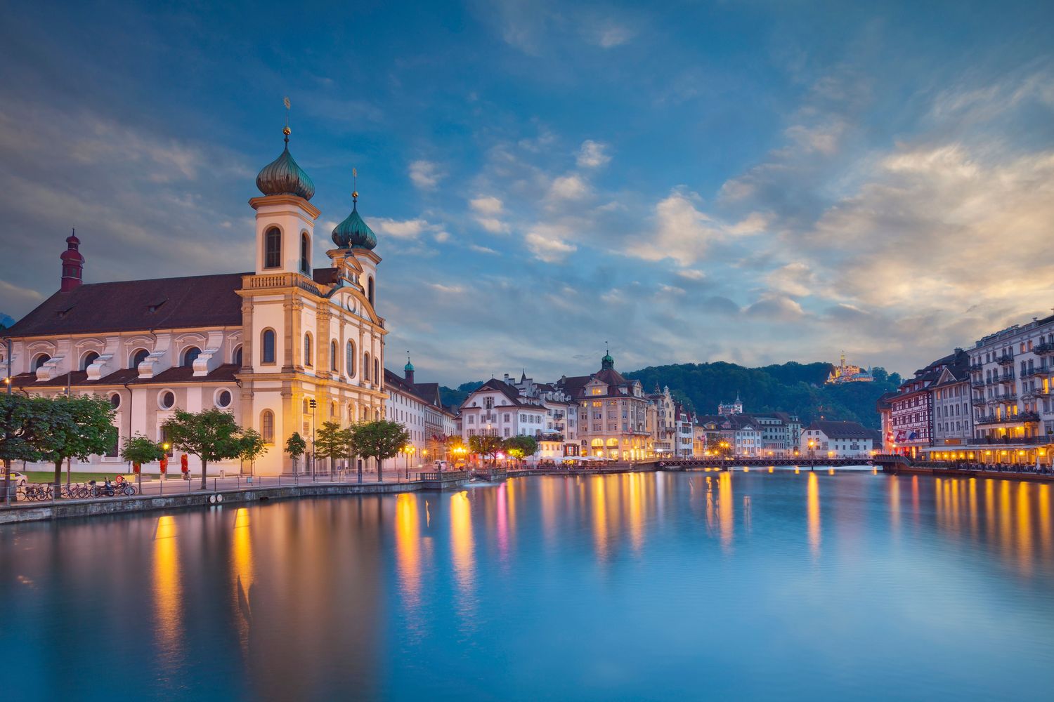 Lucerne Switzerland cityscape at dusk with Jesuit Church reflecting on River Reuss under dramatic blue sky.