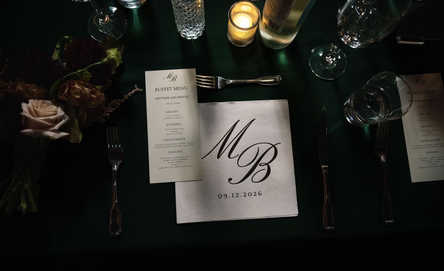 Elegant wedding table setting with monogrammed MB napkin, candles, floral centerpiece, and dark green tablecloth.