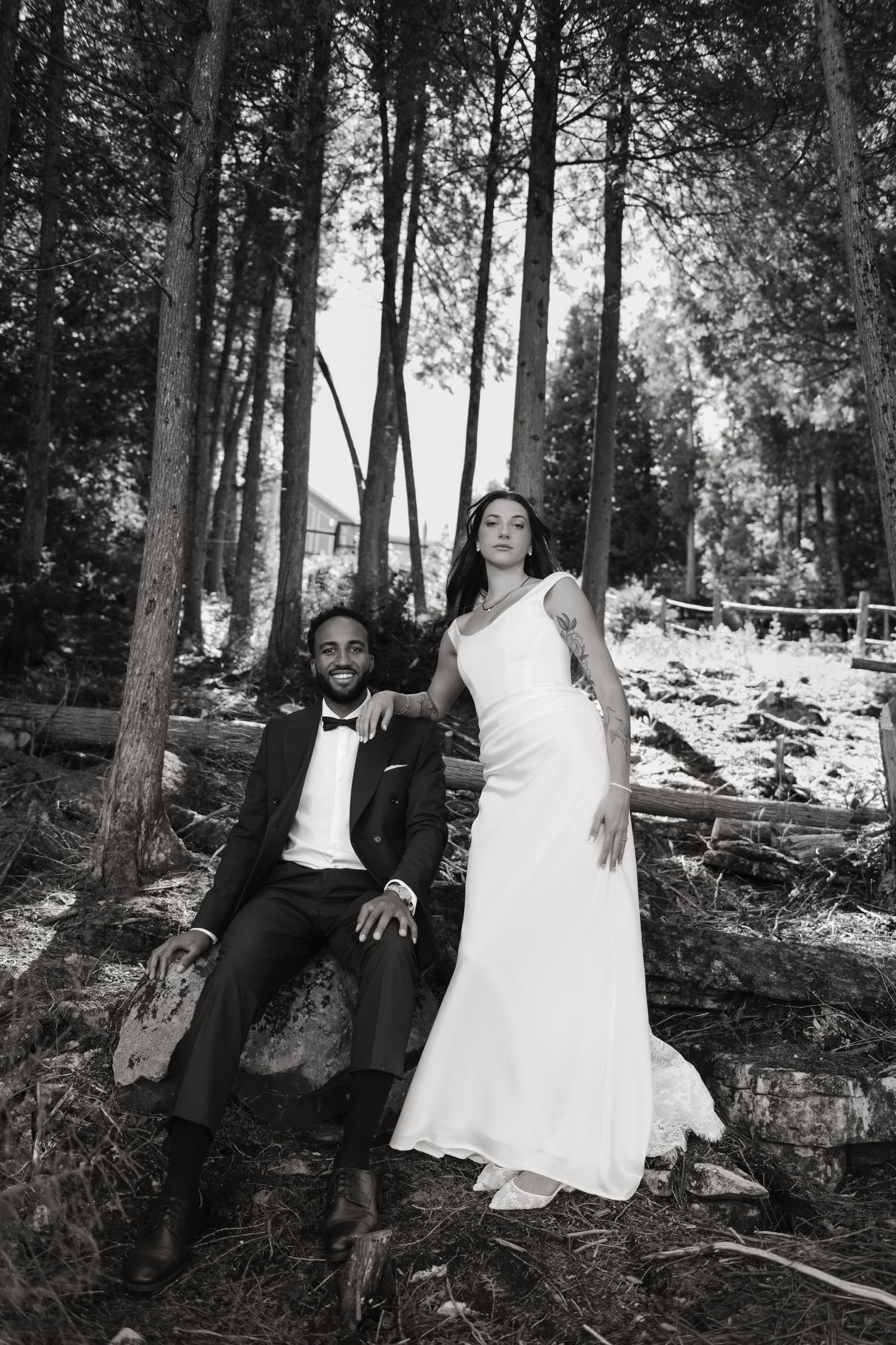 Black and white photo of bride and groom posing in a forest, groom seated on rocks in tuxedo, bride standing in white gown.