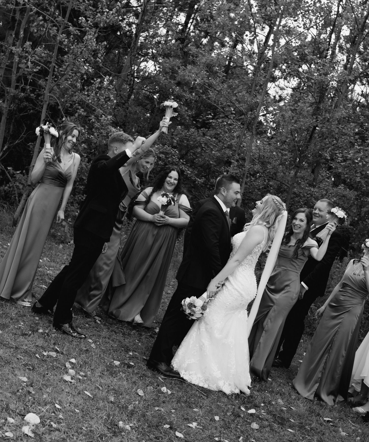 Black and white wedding party photo outdoors, bride and groom kissing surrounded by cheering bridesmaids and groomsmen.