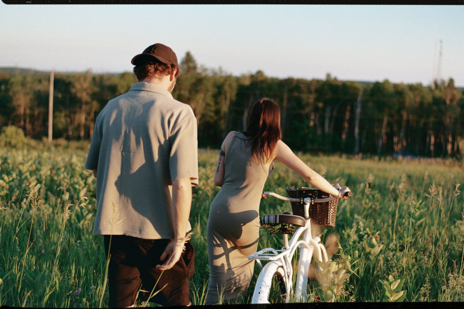 A couple walks hand in hand through a sunny meadow field with tall grass and wildflowers.
