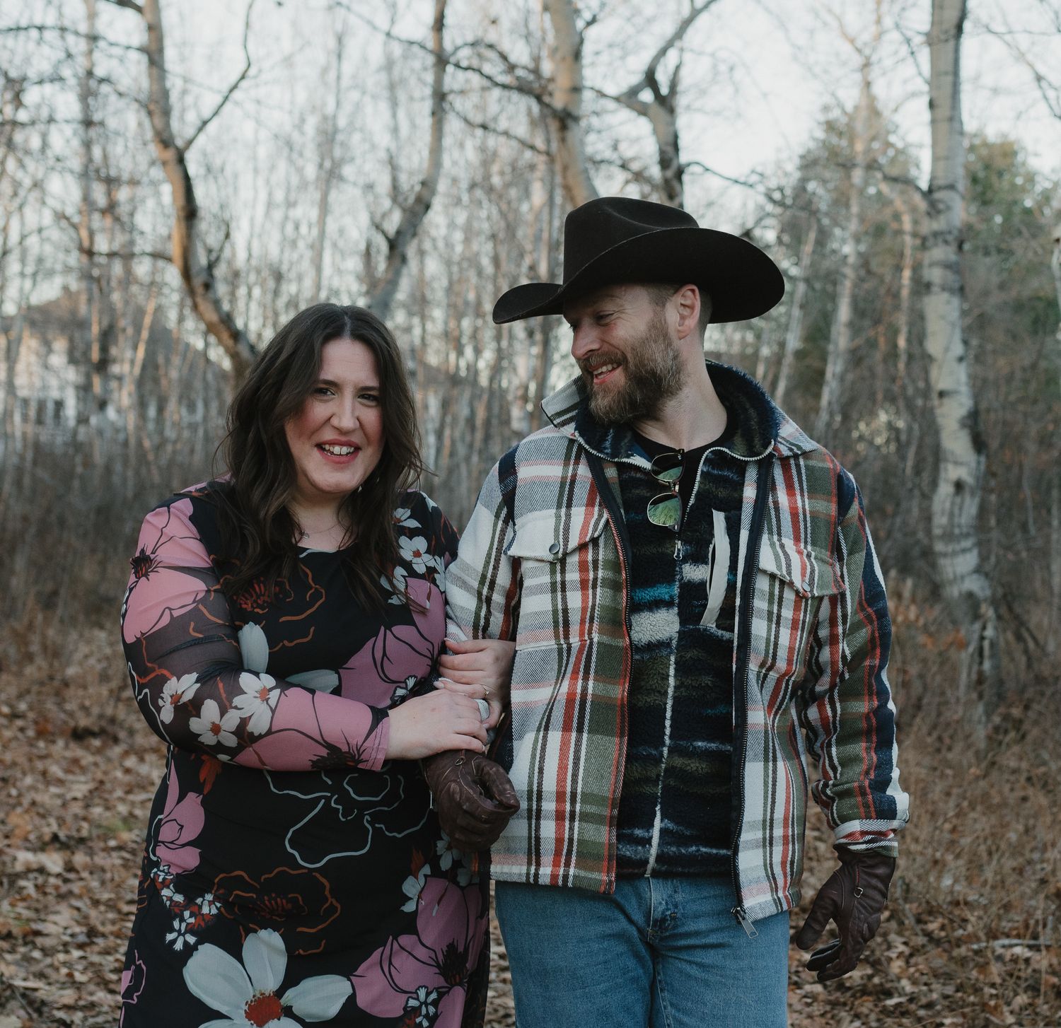 A couple shares a romantic moment in a wooded forest setting during autumn, wearing casual plaid and denim attire.