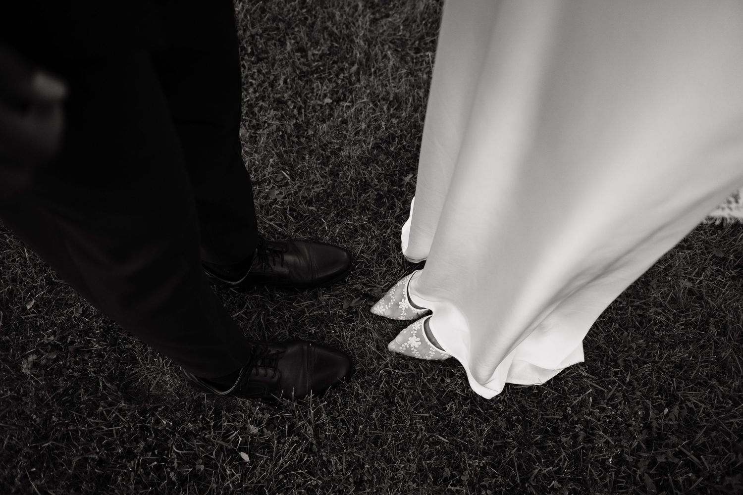 Black and white photo of bride and groom feet, wedding dress hem and dress shoes on grass.