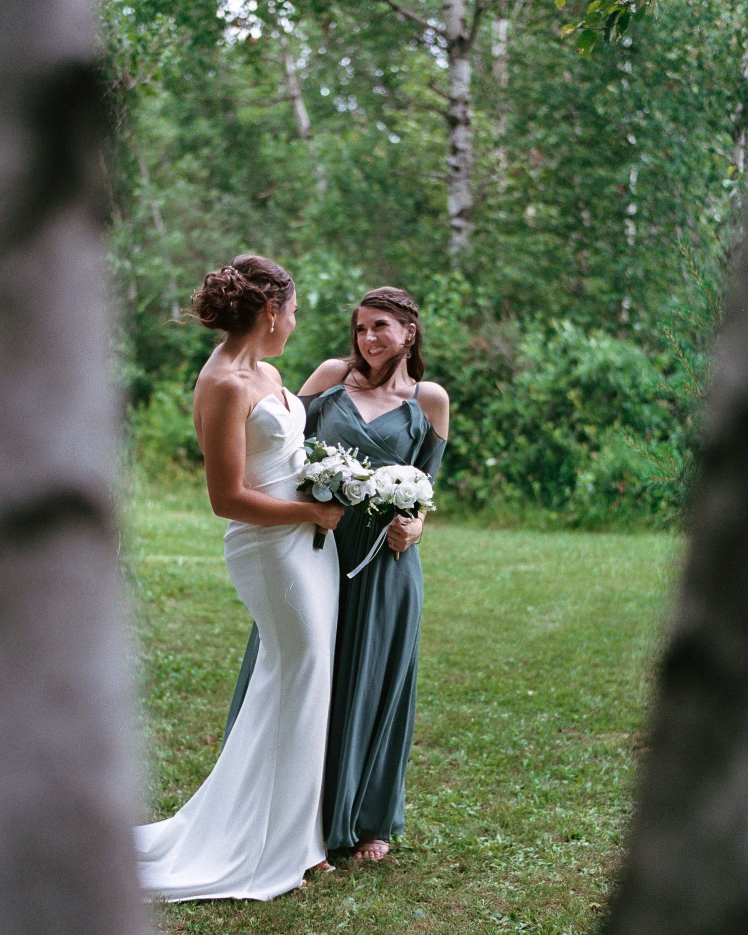 Bride in white gown and bridesmaid in sage green dress holding white bouquet smiling outdoors.