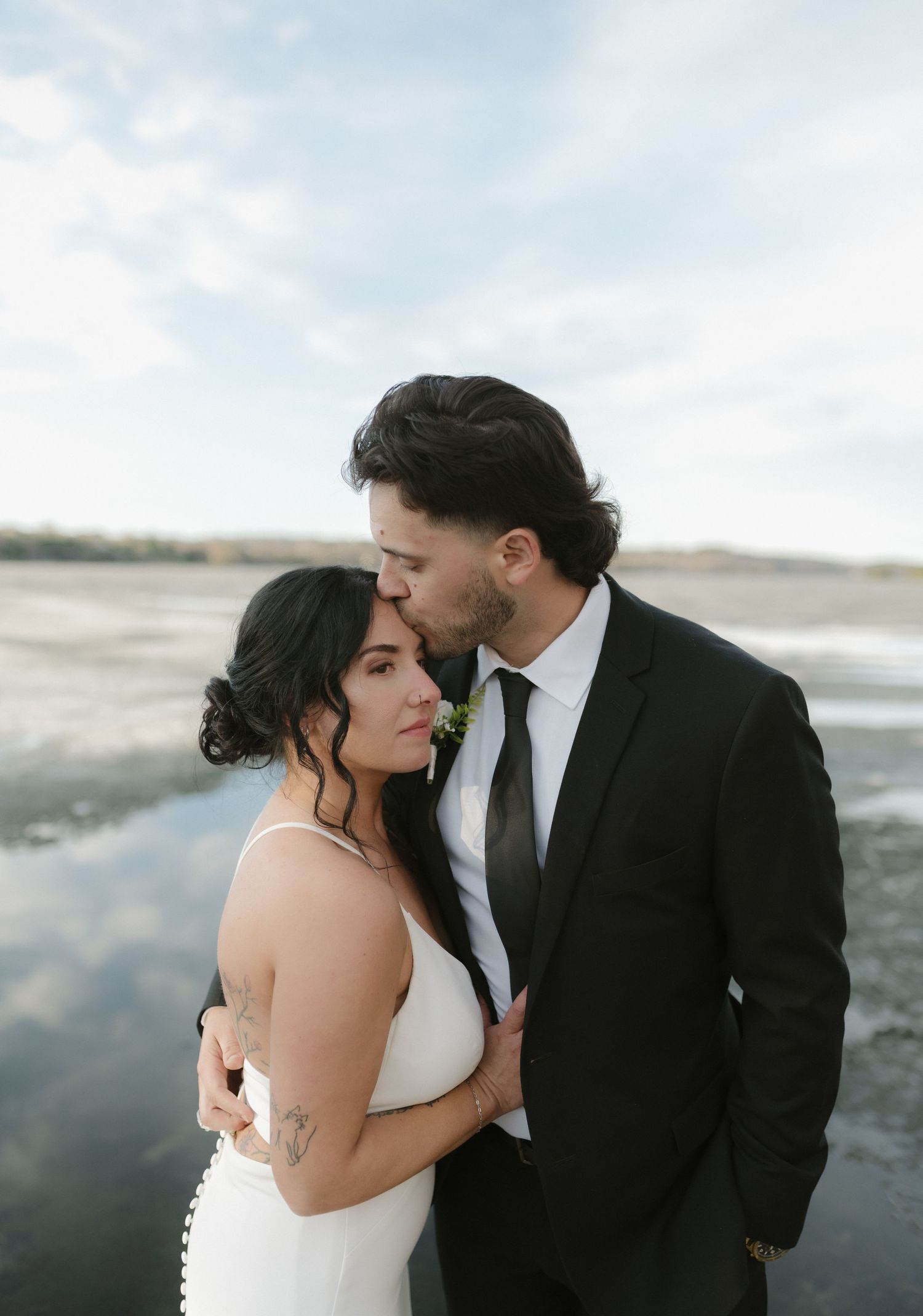 Groom in black suit kisses bride in white dress on forehead near reflective water at outdoor wedding.