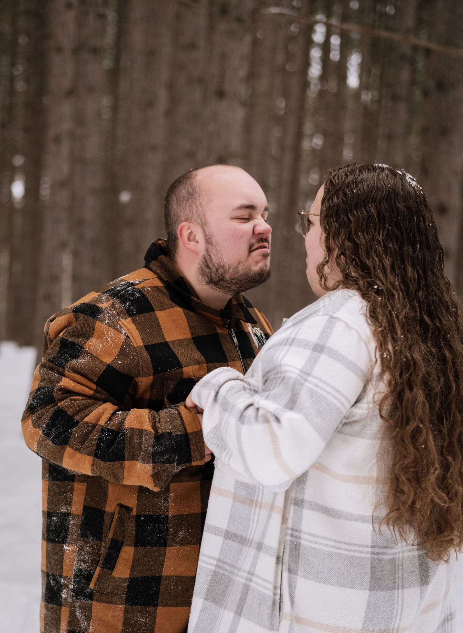 Couple sharing an intimate moment in snowy woods, man in buffalo plaid flannel and woman in white plaid jacket face each other.