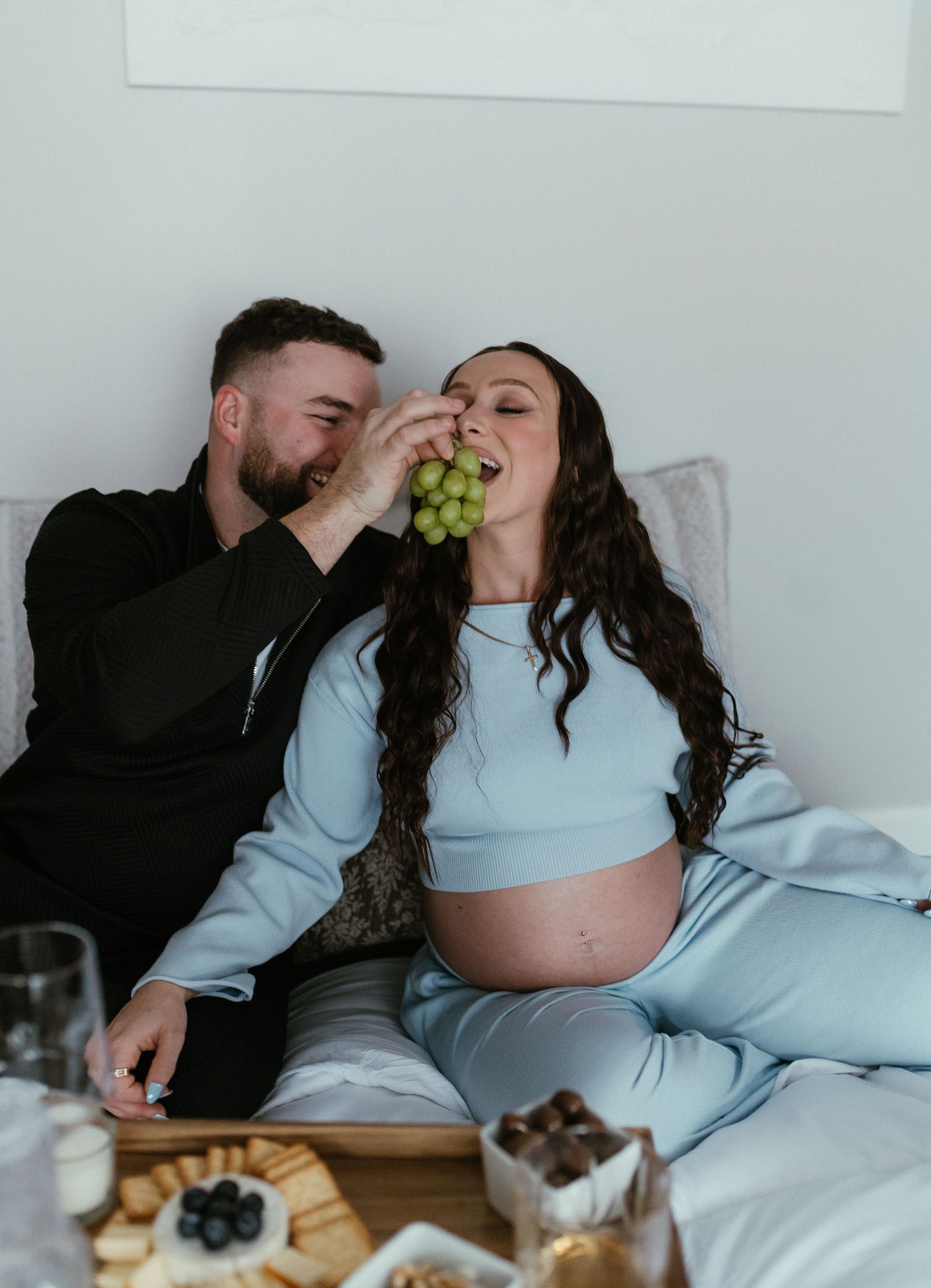 Couple sharing grapes on bed during maternity photoshoot, man feeding pregnant woman in light blue outfit.