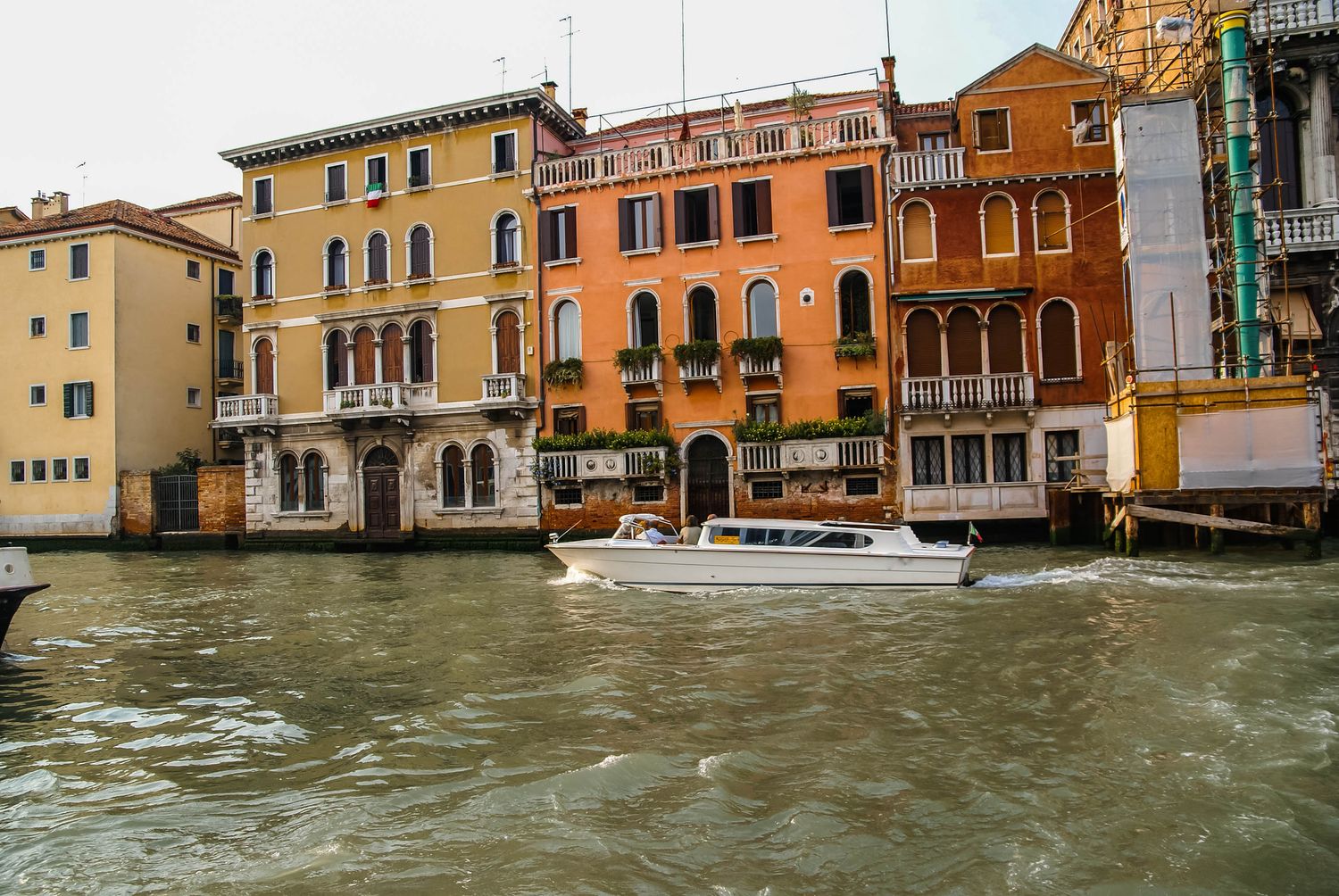 A white motorboat cruises along the Grand Canal in Venice, Italy, with colorful historic buildings lining the waterway.