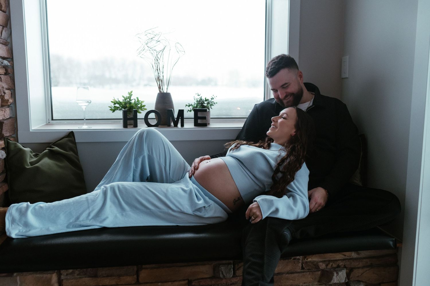 Couple sharing tender moment on couch, pregnant woman resting near window.