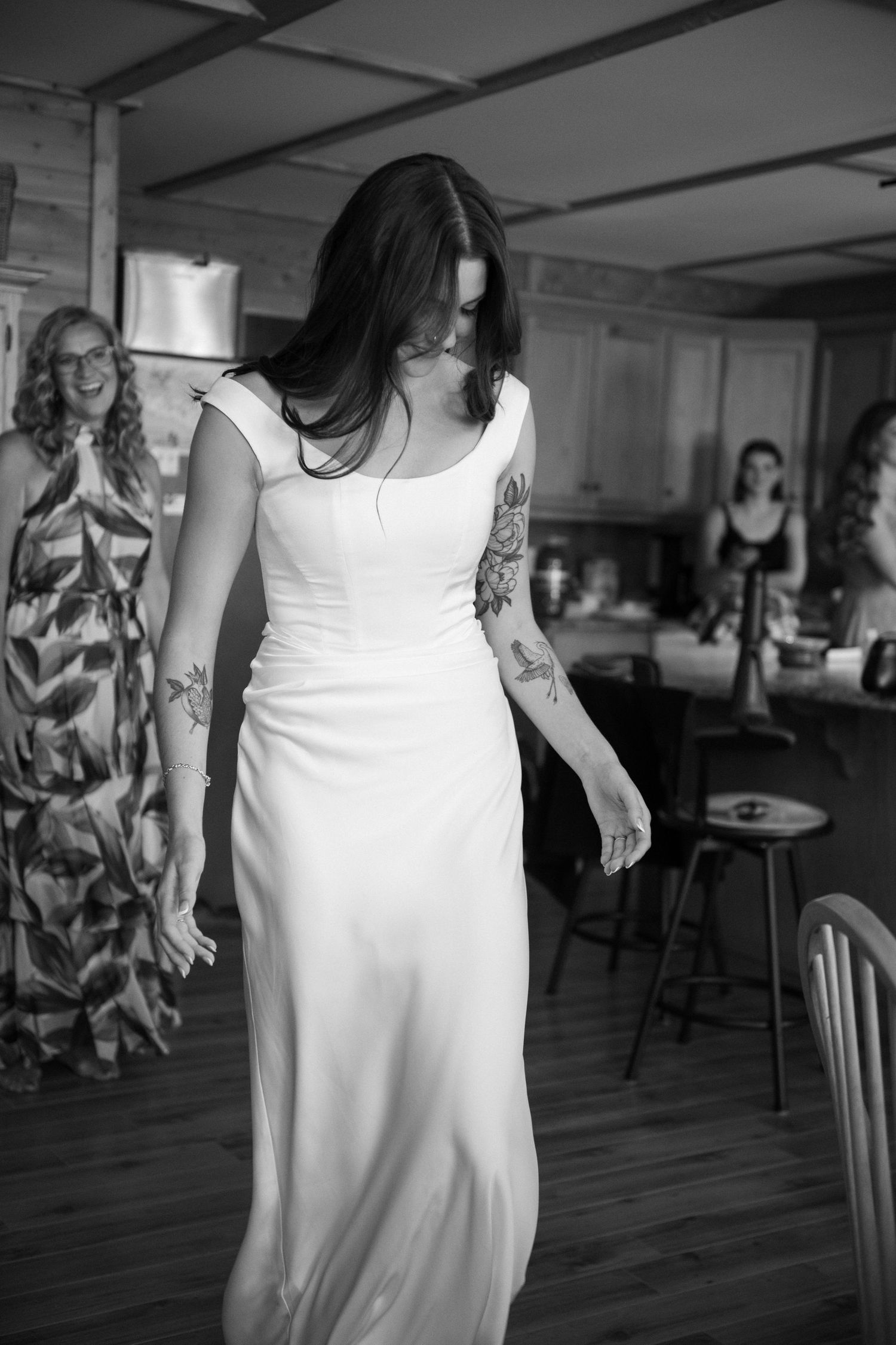 Bride in white gown with tattoos twirls while bridesmaids watch and smile in rustic indoor setting.