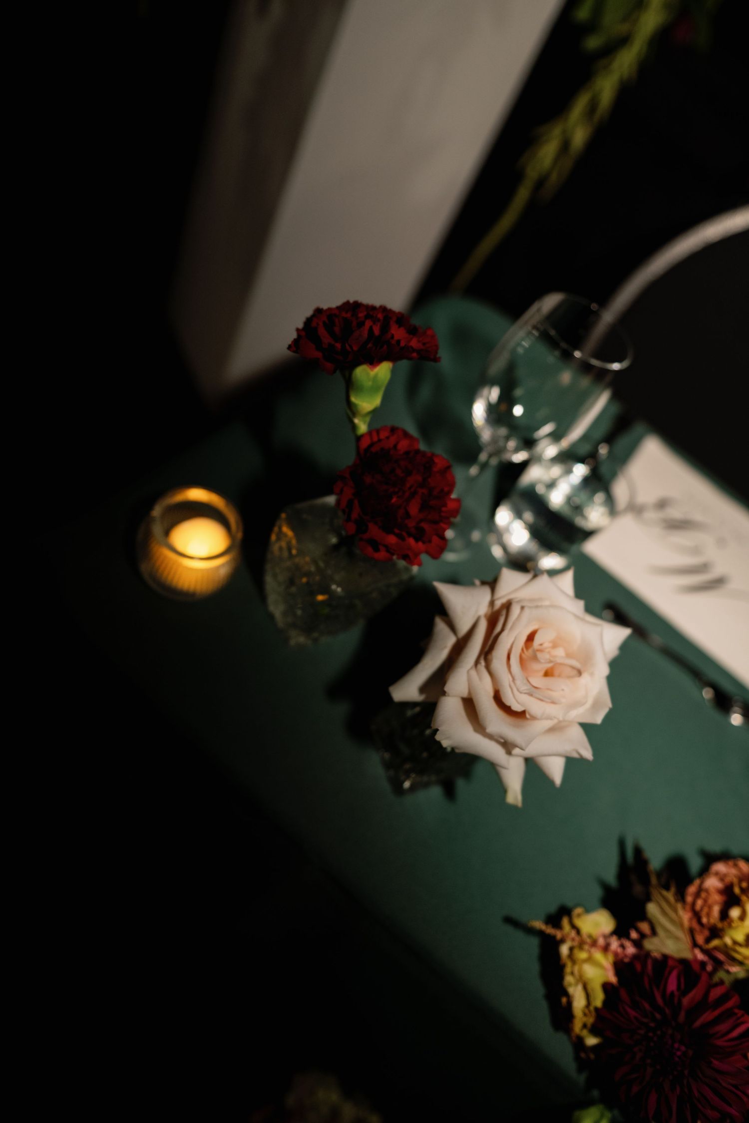 Elegant wedding table setting with white rose, red carnations, wine glass, and glowing candle on green tablecloth.